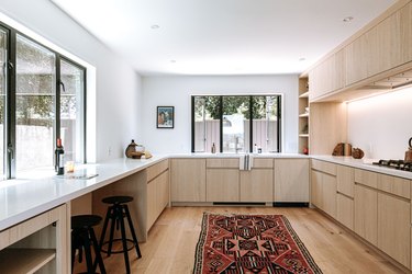 Minimalist kitchen with light wood cabinets and flooring with ornate red rug, and black frame windows